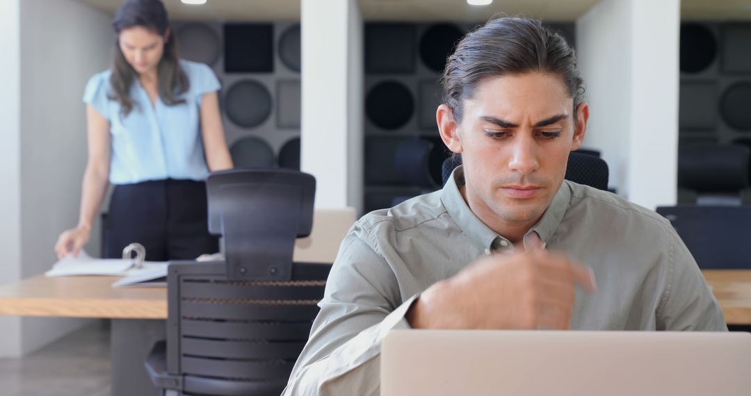 Man Working on Laptop in Office While Woman in Background Reviewing Documents - Free Images, Stock Photos and Pictures on Pikwizard.com