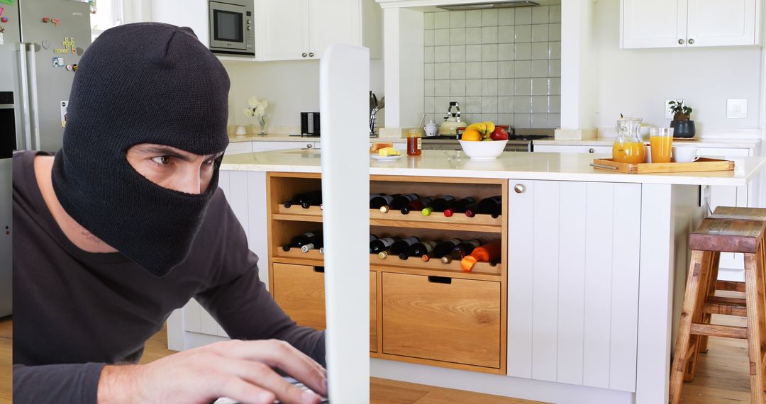 Hacker in mask typing on computer in modern kitchen