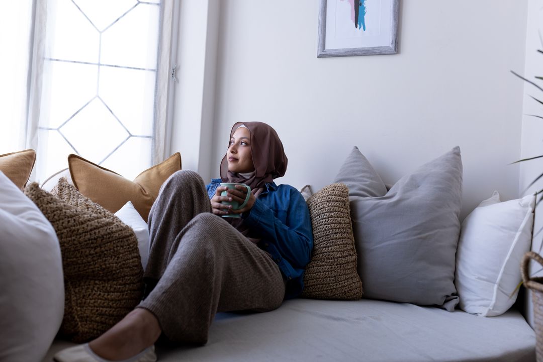 Happy biracial woman in hijab holding coffee, sitting on couch in living room at home, copy space - Free Images, Stock Photos and Pictures on Pikwizard.com