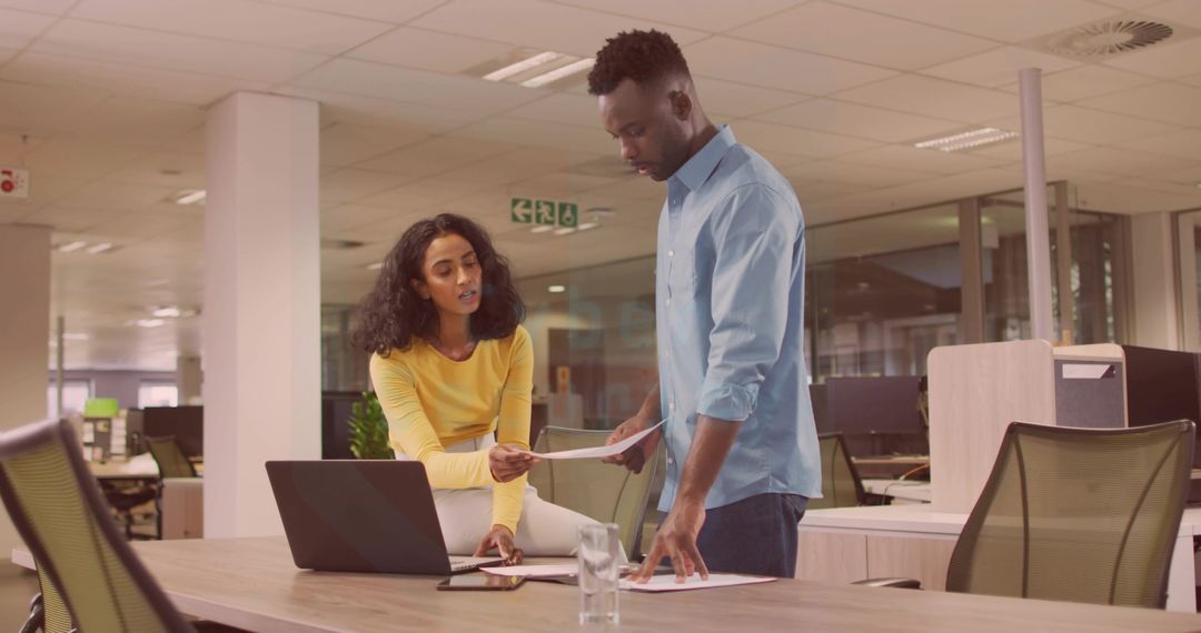 Colleagues Collaborating and Reviewing Documents at Open-Plan Office Desk with Laptop