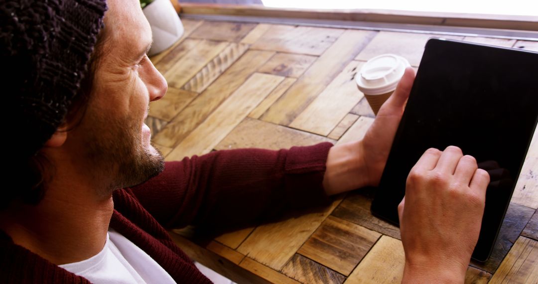 Man Using Tablet in Cafe, Wearing Beanie and Sweater, Holding Coffee Cup - Free Images, Stock Photos and Pictures on Pikwizard.com