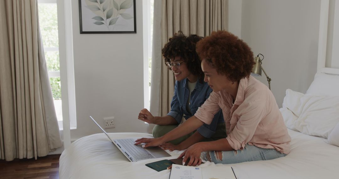 Friends planning trip on laptop while sitting on bed with passport and travel documents