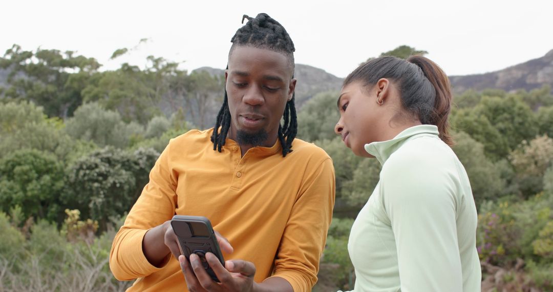 Diverse couple checking smartphone while hiking on hillside with shrubs and cloudy sky