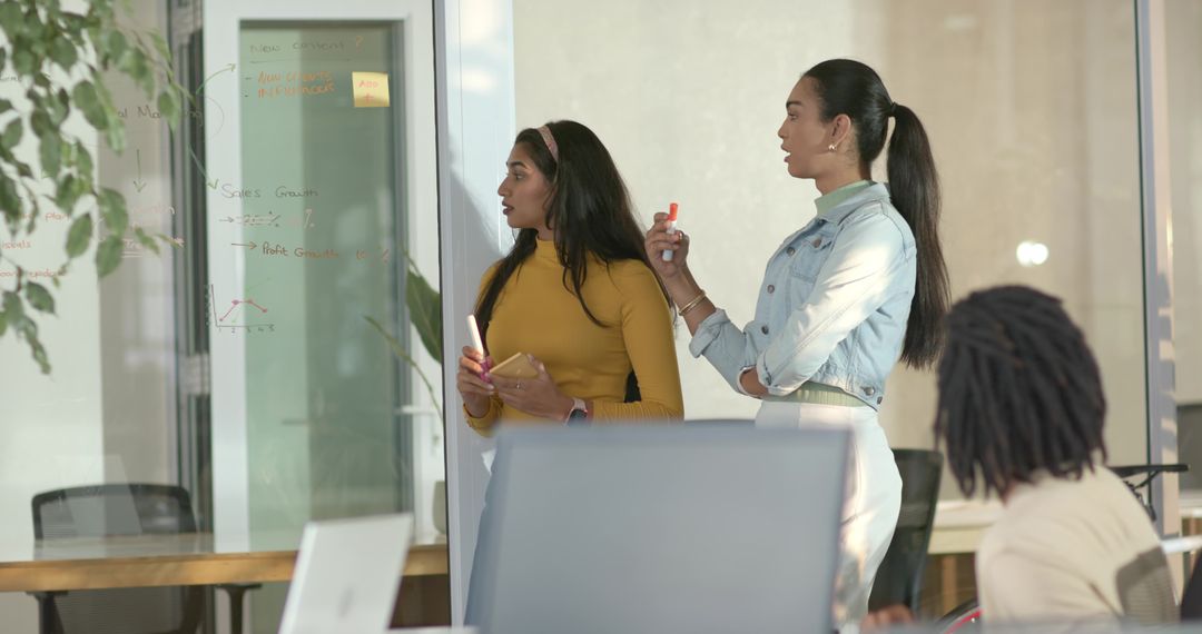 Two women collaborating at glass wall brainstorming with marker and notebook, colleague watching