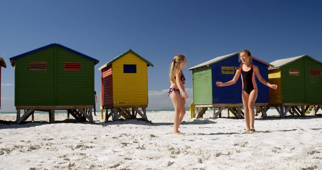 Happy caucasian girls in swimsuits standing and talking near wooden houses on sunny beach - Free Images, Stock Photos and Pictures on Pikwizard.com