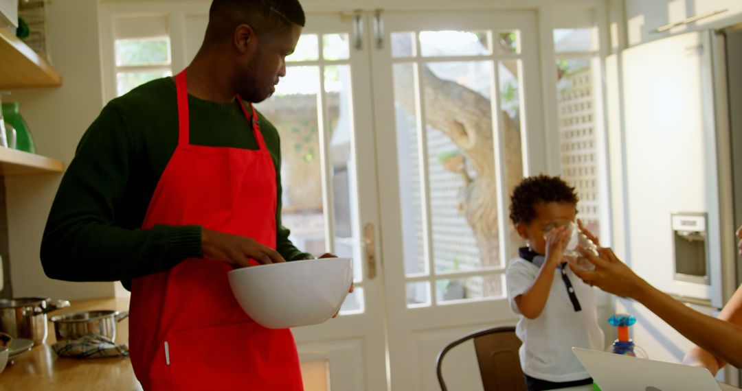 Father wearing red apron cooking while child drinks in kitchen - Free Images, Stock Photos and Pictures on Pikwizard.com