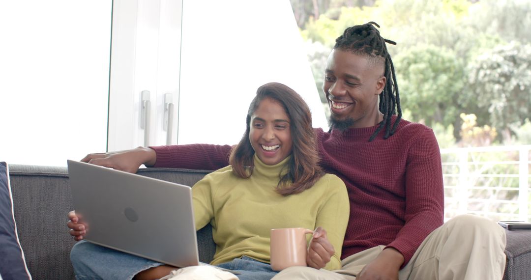 Multicultural couple relaxing on sofa using laptop and sipping coffee in sunlit living room