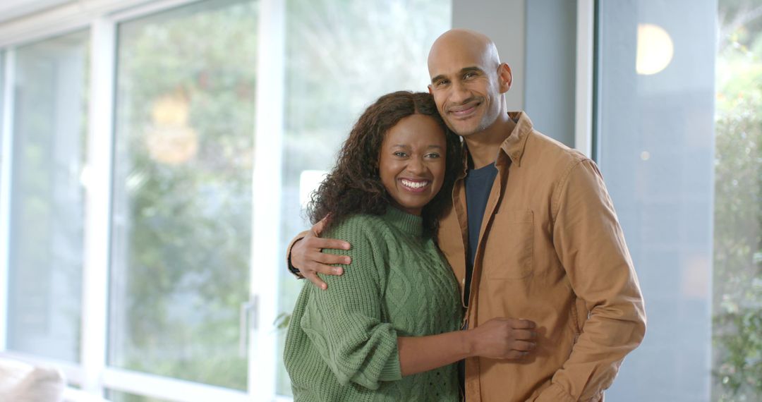 Smiling couple embracing at home by large glass doors with natural daylight and garden view