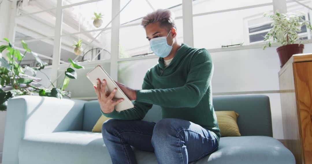 Young Man Sitting on Sofa Wearing Face Mask Using Tablet Indoors - Free Images, Stock Photos and Pictures on Pikwizard.com