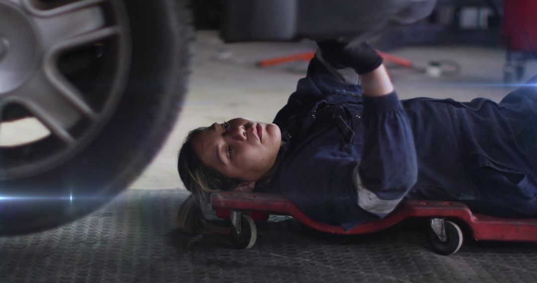 Female mechanic lying on creeper inspecting car undercarriage in garage workshop