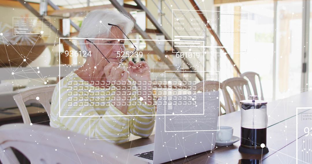 Senior woman adjusting eyeglasses while using laptop at dining table with digital AR overlay