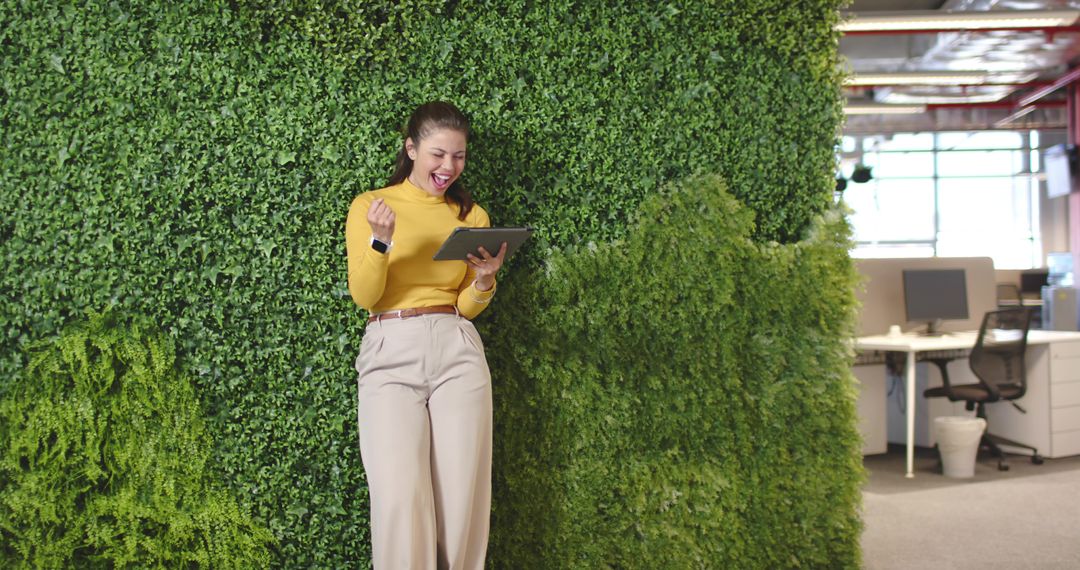 Professional celebrating while using tablet and leaning on green wall in coworking office