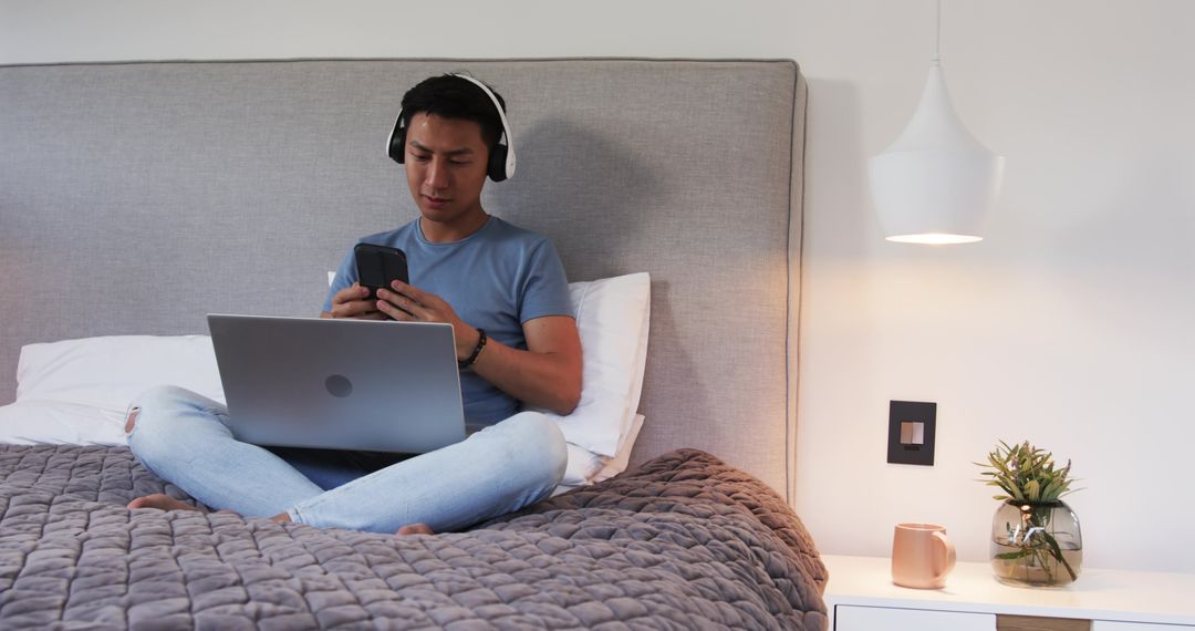 Man Using Smartphone and Laptop Seated on Bed with Headphones for Modern Relaxation