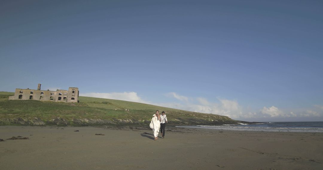 Couple Walking on Beach Near Old Stone Building Under Clear Blue Sky - Free Images, Stock Photos and Pictures on Pikwizard.com