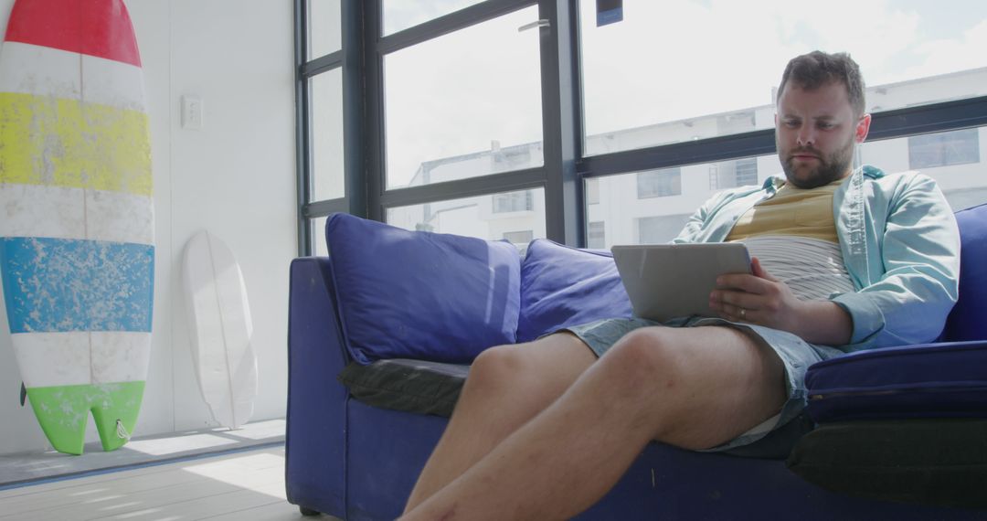 Man Using Tablet on Blue Sofa in Bright Room with Surfboard in Background - Free Images, Stock Photos and Pictures on Pikwizard.com