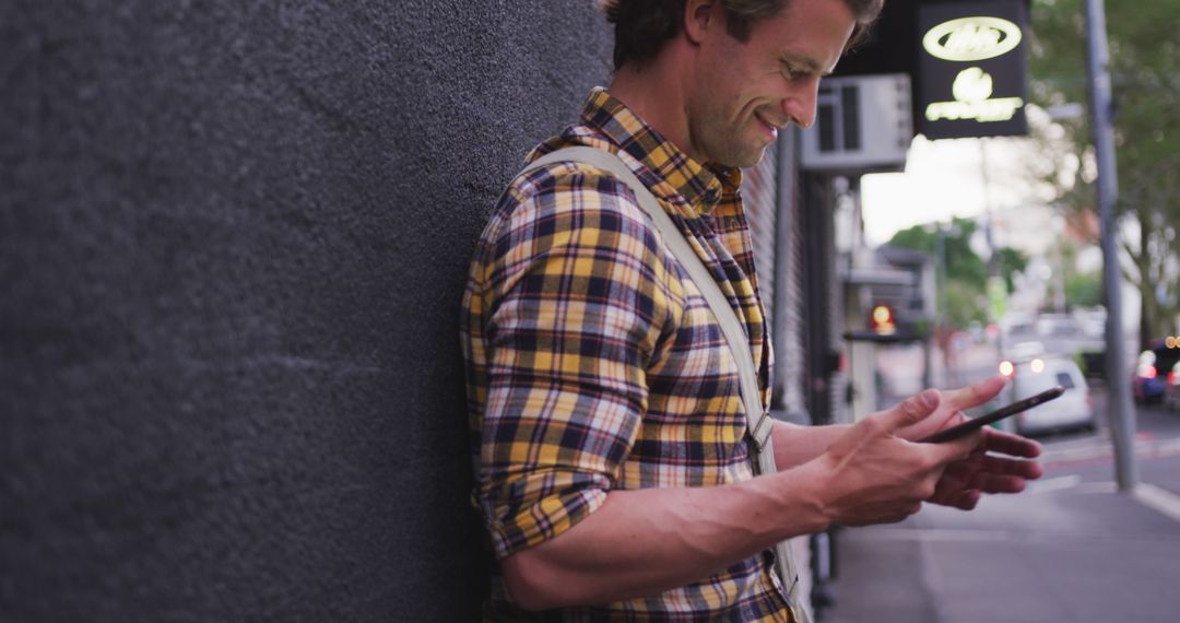 Smiling Man Leaning Against Wall Using Smartphone on City Sidewalk - Free Images, Stock Photos and Pictures on Pikwizard.com