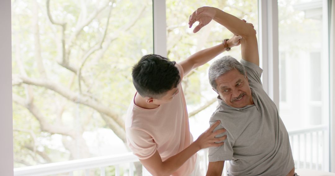 Young Man Assisting Senior with Arm Stretch Near Window - Free Images, Stock Photos and Pictures on Pikwizard.com