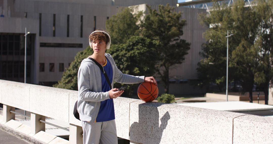 Teen male listening to music holding basketball on urban campus overpass with backpack