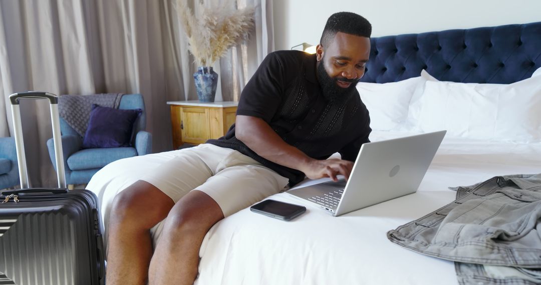 Man Typing on Laptop in Hotel Room with Suitcase Beside Him
