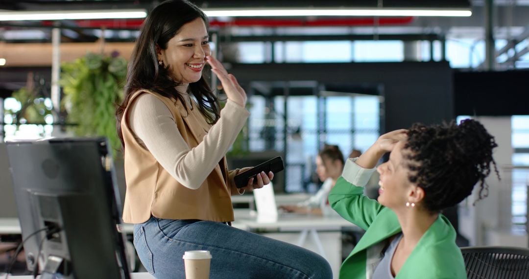 Smiling diverse female coworkers greeting and collaborating in open office with smartphone