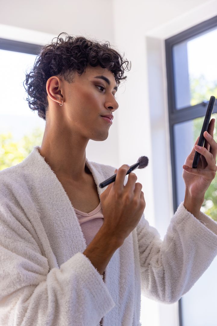 Man in Bathrobe Applying Makeup and Using Smartphone in Modern Setting
