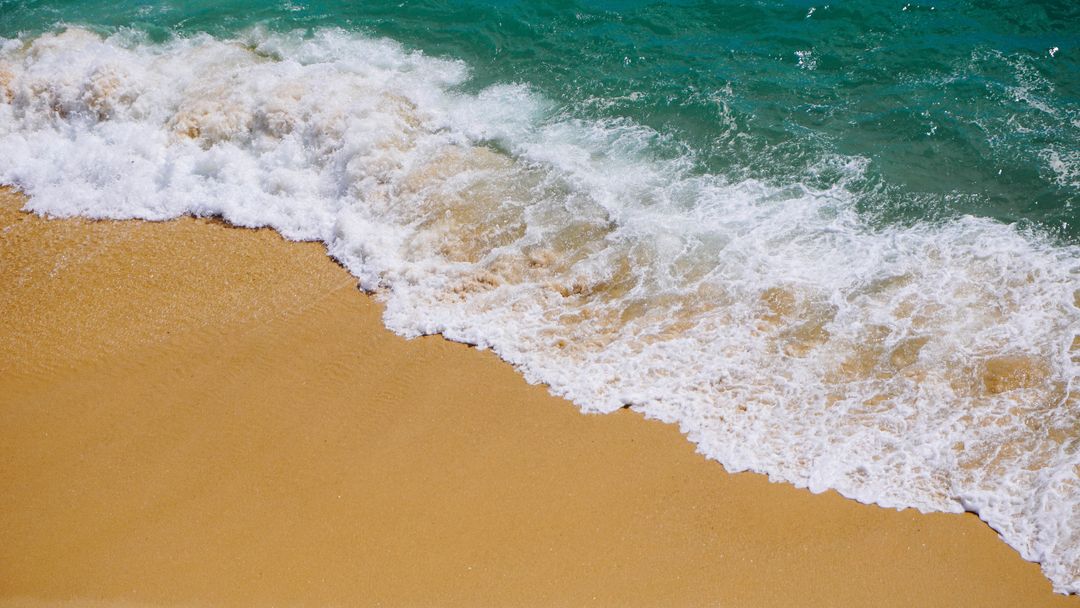 Turquoise waves crashing onto golden sand beach from above with white foam and texture