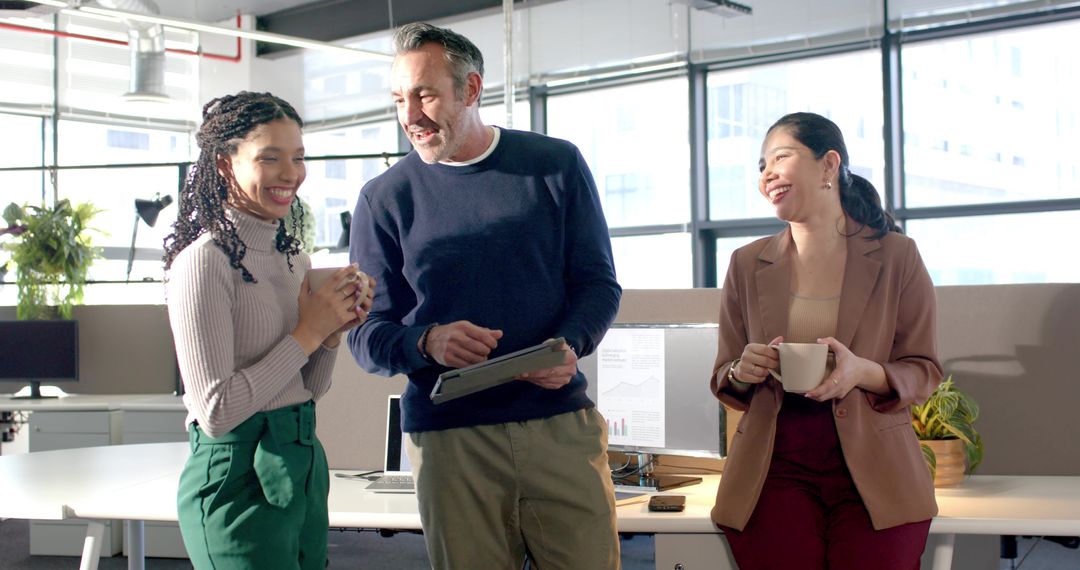 Smiling diverse coworkers sharing ideas on tablet while holding coffee in modern open-plan office