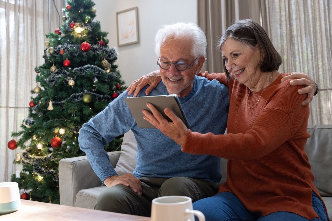 Senior Couple Sitting on Sofa Using Tablet Near Christmas Tree