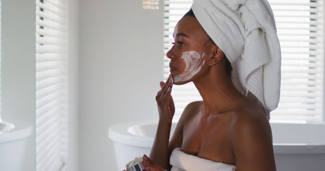 Woman Applying Facial Cream with Towel Wrapped Around Hair in Modern Bathroom - Free Images, Stock Photos and Pictures on Pikwizard.com