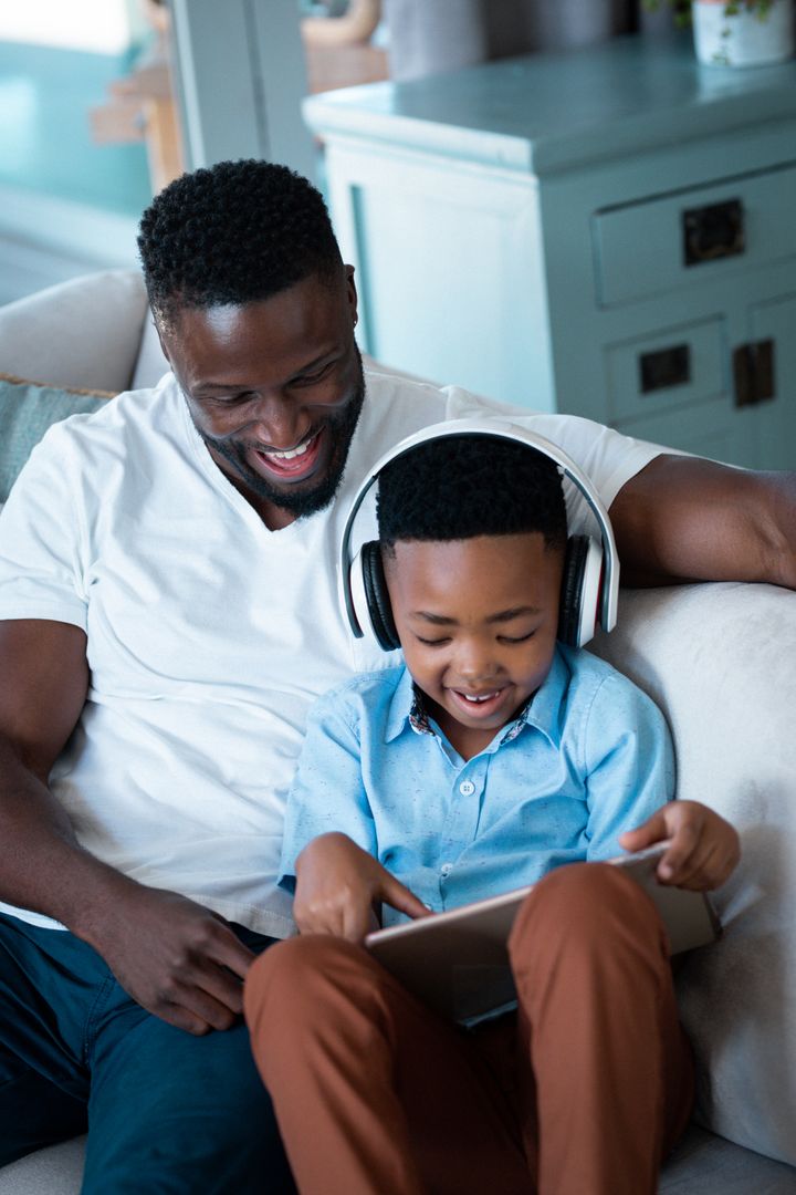African american father and son sitting on sofa, wearing headphones and using tablet - Free Images, Stock Photos and Pictures on Pikwizard.com