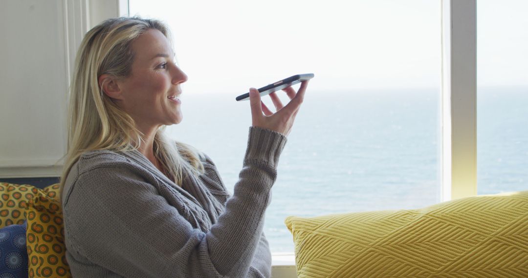 Woman Using Voice Command on Smartphone by Window Overlooking Ocean - Free Images, Stock Photos and Pictures on Pikwizard.com