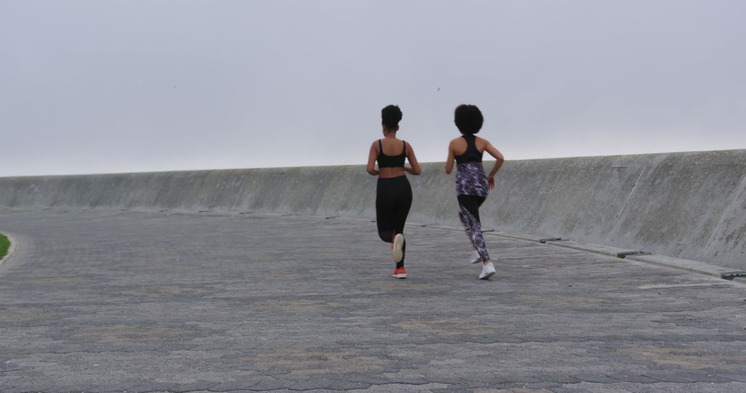 Two Women Running on Coastal Pathway on Cloudy Morning - Free Images, Stock Photos and Pictures on Pikwizard.com