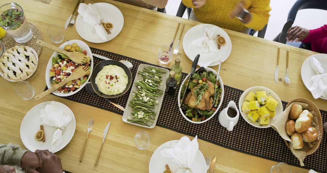 Top view of diverse family sharing roasted chicken, salad, corn and rolls on wooden table
