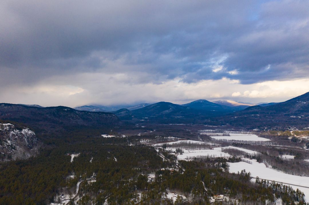 Winter mountain valley showing snow-covered fields and pine forest under dramatic clouds