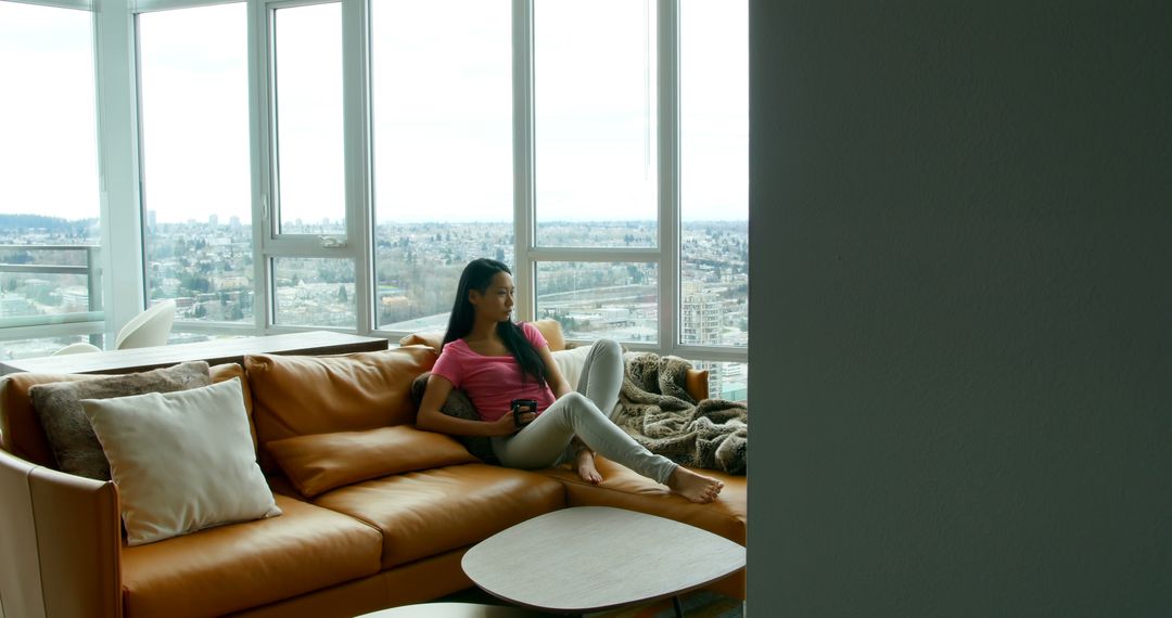 Young woman relaxing on sofa in modern high-rise apartment with city view - Free Images, Stock Photos and Pictures on Pikwizard.com