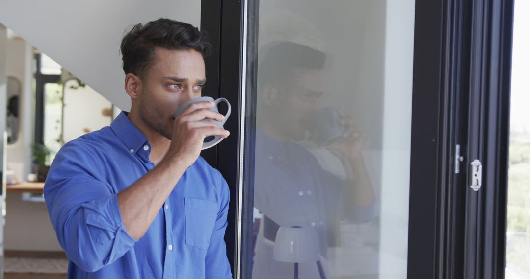 Young man in blue shirt drinking coffee and looking outside - Free Images, Stock Photos and Pictures on Pikwizard.com