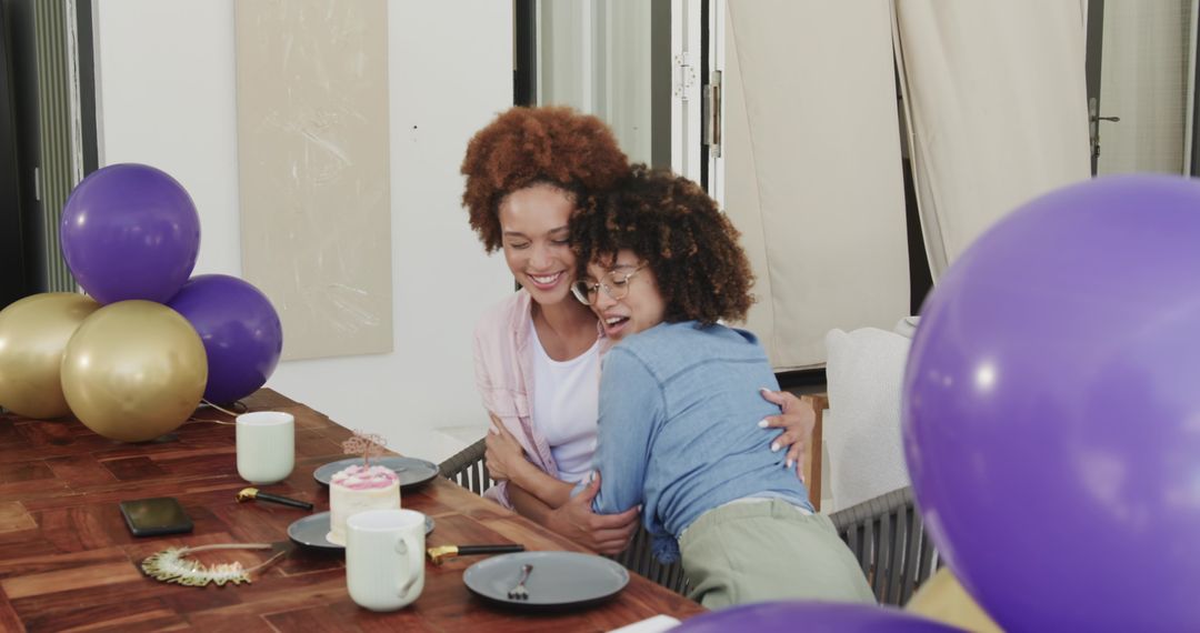 Two diverse female friends hugging and smiling at birthday brunch with purple balloons and cake