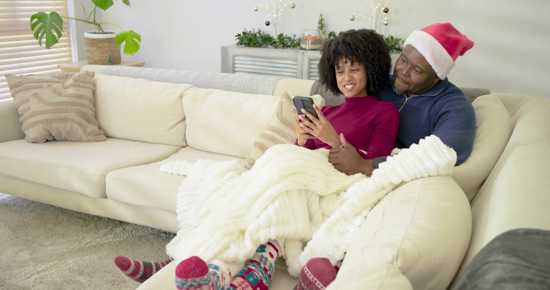 Couple cuddling on sofa under chunky blanket, scrolling smartphone, wearing Santa hat