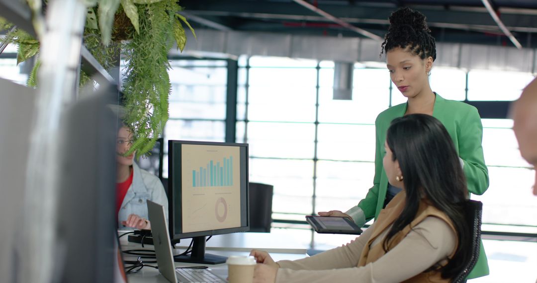Collaborative team analyzing charts on monitor and tablet in sunlit open-plan office