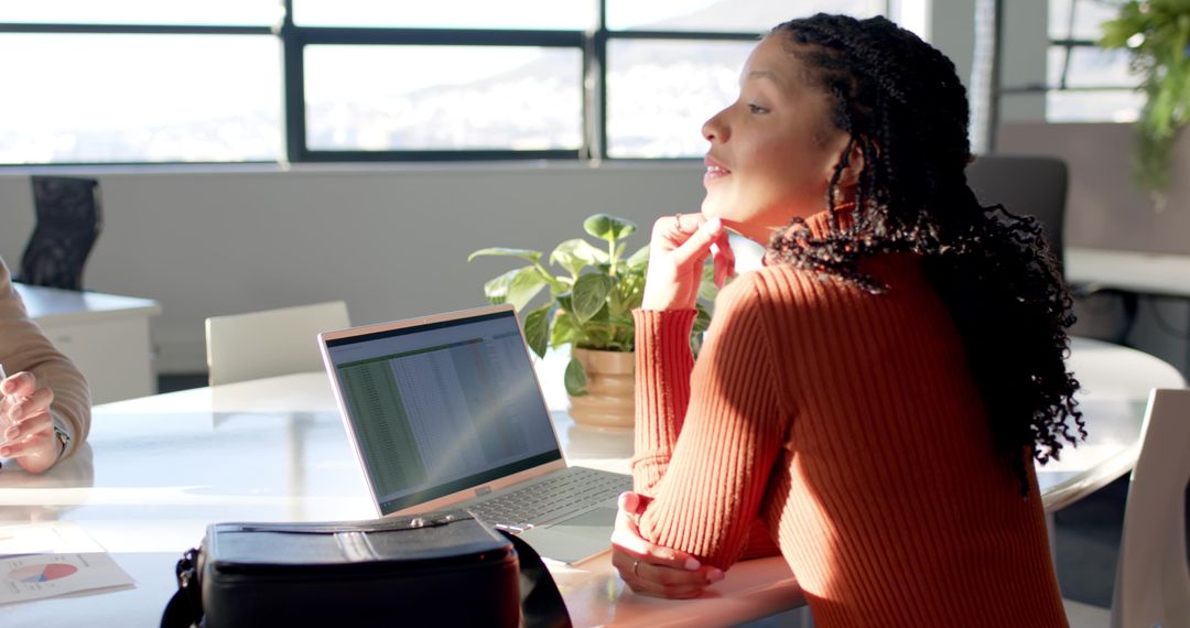 Diverse female colleague leaning and collaborating at sunlit modern office table with laptop