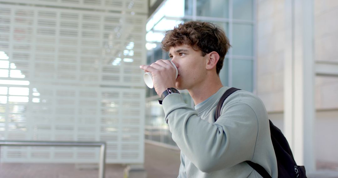 Young man drinking takeaway coffee on modern campus walkway with backpack