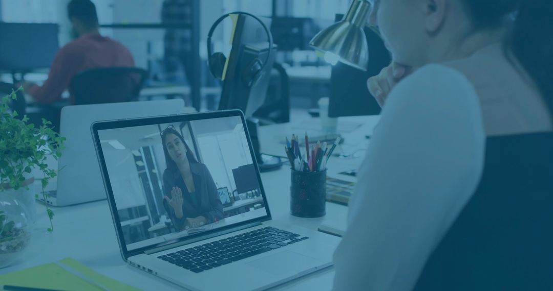 Woman joining remote video call in open-plan office, working at laptop with pens
