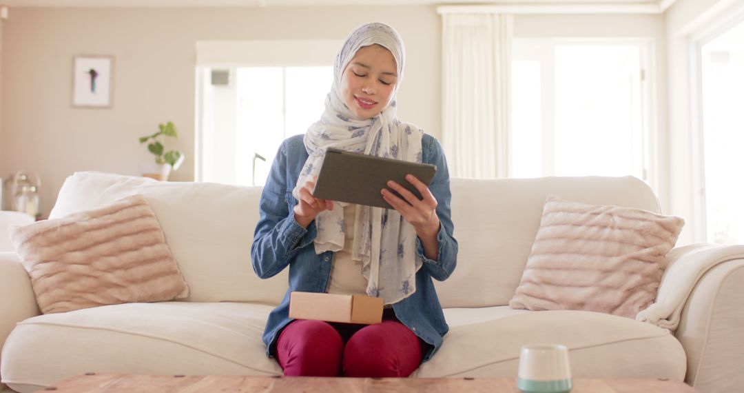 Muslim Woman Opening Parcel with Tablet in Comfortable Home Setting - Free Images, Stock Photos and Pictures on Pikwizard.com