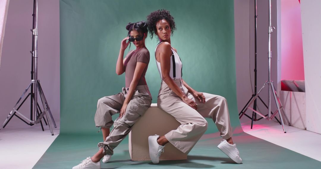 Two models sitting back-to-back posing in studio wearing cargo pants and white sneakers