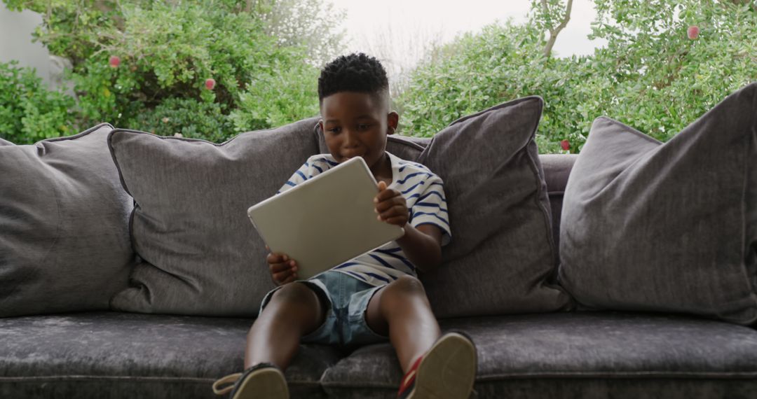 Young African American Boy Relaxing On Sofa Using Tablet - Free Images, Stock Photos and Pictures on Pikwizard.com