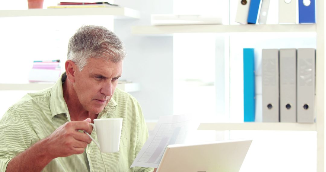 Middle Aged Man Drinking Coffee While Reading Documents on Laptop - Free Images, Stock Photos and Pictures on Pikwizard.com