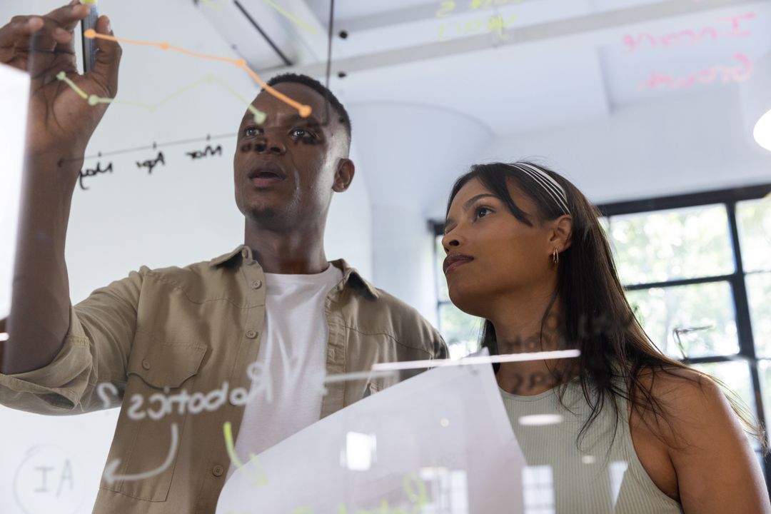 Diverse coworkers collaborating and analyzing charts on glass board during brainstorming