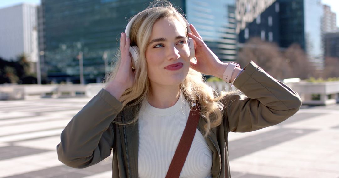 Blonde woman listening to music with wireless over-ear headphones in sunlit city plaza