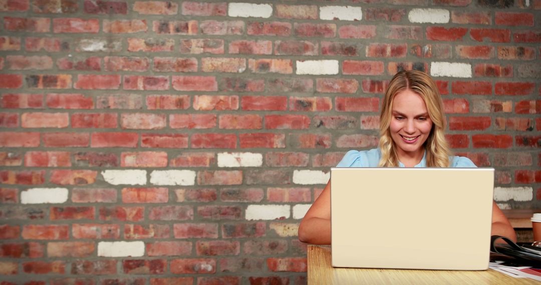 Smiling Woman Working on Laptop in Office with Brick Wall Background - Free Images, Stock Photos and Pictures on Pikwizard.com