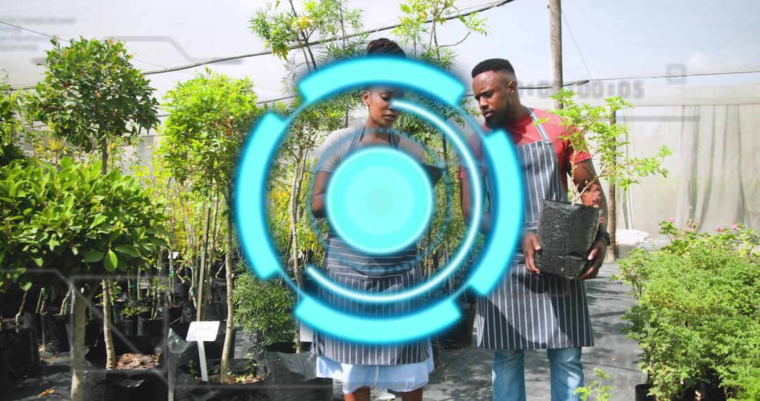 Two Nursery Workers Consulting Notepad While Holding Sapling in Black Pot in Greenhouse